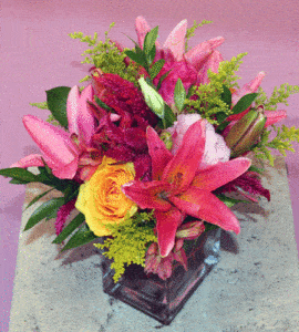 A gorgeous assorment of colorful Lilies, Roses, Alstromeria, and various other assorted flowers, tastefully arranged in an elegant, heavy glass vase.