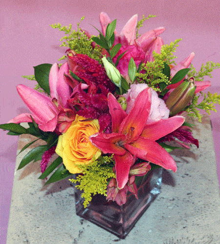 A gorgeous assorment of colorful Lilies, Roses, Alstromeria, and various other assorted flowers, tastefully arranged in an elegant, heavy glass vase.